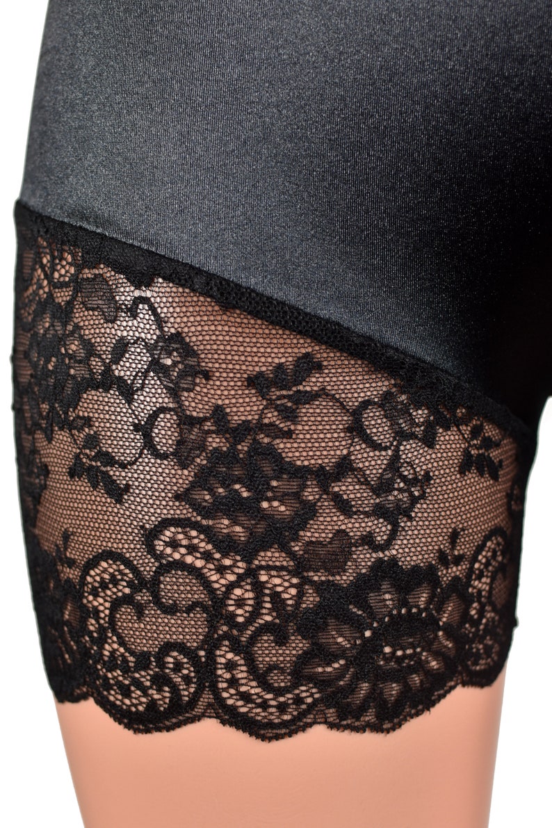 May include: A pair of black shorts featuring a smooth, slightly reflective fabric upper section and a wide, intricate black floral lace trim along the bottom edge. The lace has a scalloped hemline and a detailed, open-work botanical pattern.