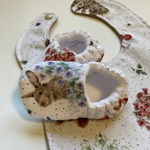 May include: A set of baby booties and a bib with a white background and a floral pattern featuring a rabbit, a hedgehog, and a robin. The booties and bib are made of soft, comfortable fabric.