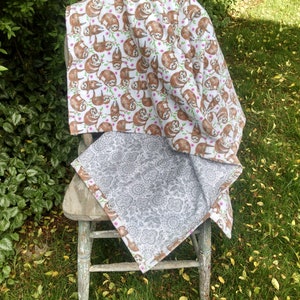 May include: A white and gray reversible blanket with a sloth pattern on one side and a floral pattern on the other side. The blanket is draped over a wooden chair in a grassy area.