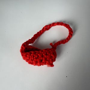 Puede incluir: Peque&ntilde;o bolso rojo hecho a mano con ganchillo, con un asa en forma de bucle. El bolso est&aacute; hecho de hilo grueso y tiene un aspecto texturizado. El asa tambi&eacute;n est&aacute; tejida a ganchillo y fijada a la parte superior del bolso.