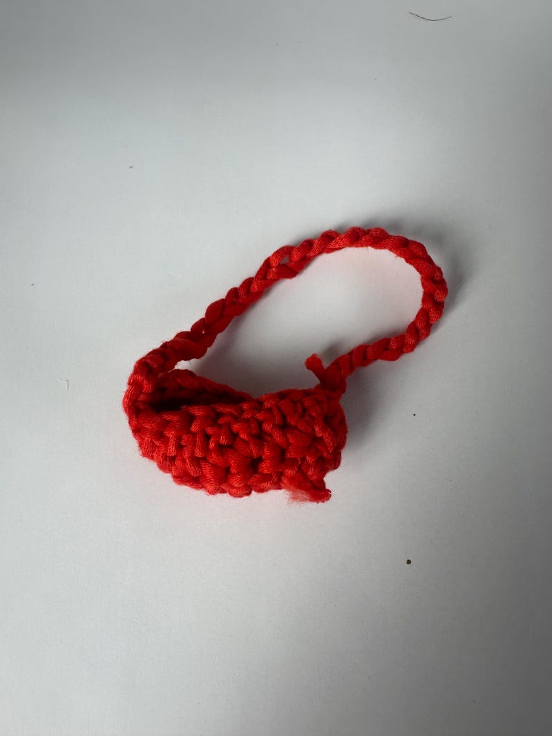 Puede incluir: Peque&ntilde;o bolso rojo hecho a mano con ganchillo, con un asa en forma de bucle. El bolso est&aacute; hecho de hilo grueso y tiene un aspecto texturizado. El asa tambi&eacute;n est&aacute; tejida a ganchillo y fijada a la parte superior del bolso.