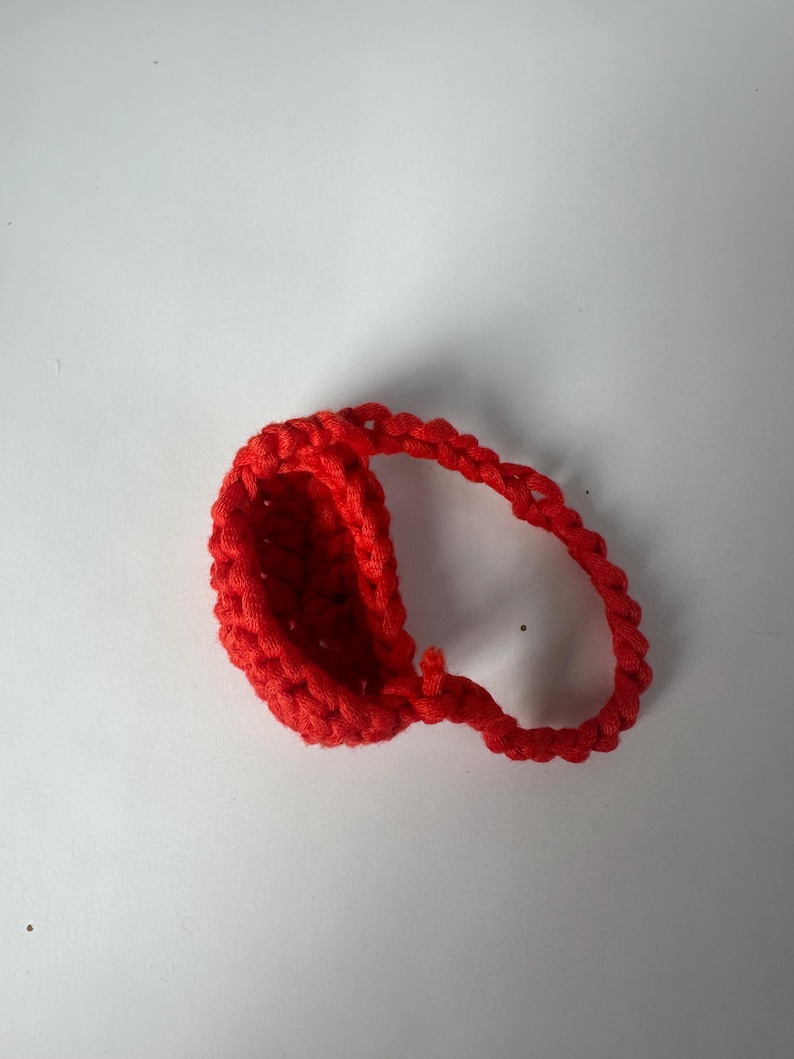 Puede incluir: Un art&iacute;culo de ganchillo rojo brillante hecho a mano, posiblemente una peque&ntilde;a cesta o contenedor. El art&iacute;culo est&aacute; hecho de hilo grueso y tiene una apariencia texturizada. La forma es ovalada con un asa en bucle. El fondo es blanco liso.
