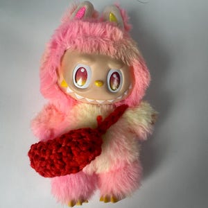 Puede incluir: Un juguete de peluche con un cuerpo peludo rosa y amarillo, que lleva un atuendo con capucha rosa con orejas. El juguete tiene una cara sonriente con ojos grandes y un accesorio rojo de ganchillo.