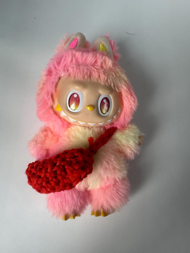 Puede incluir: Un juguete de peluche con un cuerpo peludo rosa y amarillo, que lleva un atuendo con capucha rosa con orejas. El juguete tiene una cara sonriente con ojos grandes y un accesorio rojo de ganchillo.