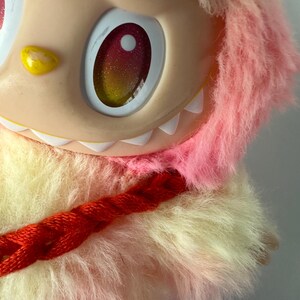 Puede incluir: Primer plano de un juguete de peluche con una cara melocot&oacute;n claro, ojos grandes con iris rojos y amarillos brillantes y una hilera de dientes blancos. El juguete tiene pelaje rosa y amarillo esponjoso y un detalle trenzado rojo.
