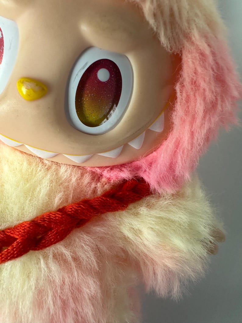 Puede incluir: Primer plano de un juguete de peluche con una cara melocot&oacute;n claro, ojos grandes con iris rojos y amarillos brillantes y una hilera de dientes blancos. El juguete tiene pelaje rosa y amarillo esponjoso y un detalle trenzado rojo.