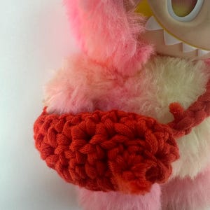 Puede incluir: Un juguete de peluche con un cuerpo peludo rosa y amarillo, una envoltura de brazo de ganchillo roja y una cara grande de dibujos animados con un solo ojo. El juguete tiene un dise&ntilde;o caprichoso y una textura suave.