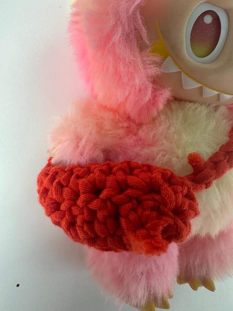 Puede incluir: Un juguete de peluche con un cuerpo peludo rosa y amarillo, una envoltura de brazo de ganchillo roja y una cara grande de dibujos animados con un solo ojo. El juguete tiene un dise&ntilde;o caprichoso y una textura suave.