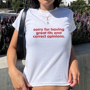 Può includere: T-shirt bianca con la scritta rossa "sorry for having great tits and correct opinions." La maglietta ha maniche corte e girocollo. La persona indossa occhiali da sole e una borsa nera.