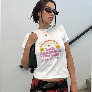 Puede incluir: Camiseta blanca con un diseño de arcoíris, sol y mariposas. El texto en la camiseta dice "LOUD AND PROUD QUEER QUEEN" en una fuente colorida y retro. La persona lleva gafas de sol.