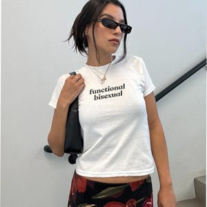 May include: A white t-shirt with the text "functional bisexual" in black. The person is wearing black sunglasses and a floral skirt with red and black flowers. A black purse is over the shoulder.