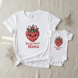 Op de afbeelding: Witte t-shirt en babybody set met een cartoon aardbei ontwerp en roze strik. Het t-shirt heeft de tekst "Berry Sweet Mama", en de body "Berry Sweet Mini". Perfect voor bijpassende outfits.