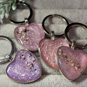 May include: A collection of heart-shaped keyrings in pink and purple hues, with silver metal rings. Each heart pendant is filled with a textured, shimmering material. The keyrings are displayed on a soft, grey surface.