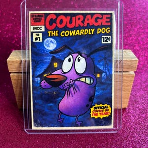 May include: A comic book cover featuring Courage the Cowardly Dog. The title "COURAGE THE COWARDLY DOG" is in red, with the character in the foreground. The comic is in a protective sleeve, with the text "OFFICIAL COMIC OF THE YEAR!"
