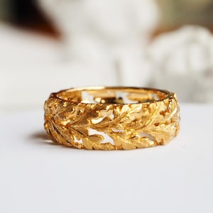 Op de afbeelding: Een goudkleurige ring met een ingewikkeld bladerenontwerp. De ring is een bandstijl, met de bladeren uitgesneden om een opengewerkt patroon te creëren. De ring staat tegen een witte achtergrond, met een vage achtergrond.