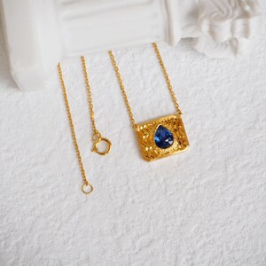 May include: A gold chain necklace with a pendant featuring a blue teardrop-shaped gemstone set in a textured gold frame.