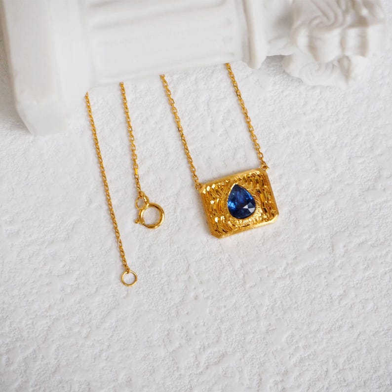 May include: A gold chain necklace with a pendant featuring a blue teardrop-shaped gemstone set in a textured gold frame.