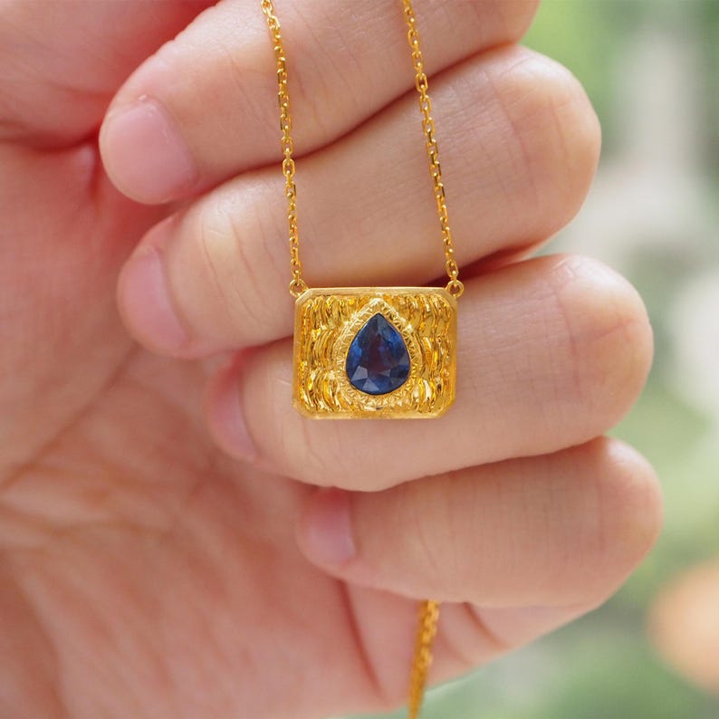 May include: A gold necklace with a rectangular pendant featuring a blue teardrop-shaped gemstone.