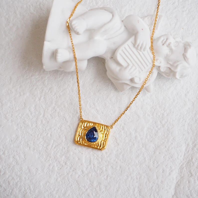 May include: A gold chain necklace with a rectangular pendant featuring a blue teardrop-shaped gemstone set in a textured gold frame.