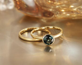 May include: Two gold rings, one with a round, dark teal gemstone. The rings are set against a warm, golden background. The rings are likely for engagement or wedding purposes.