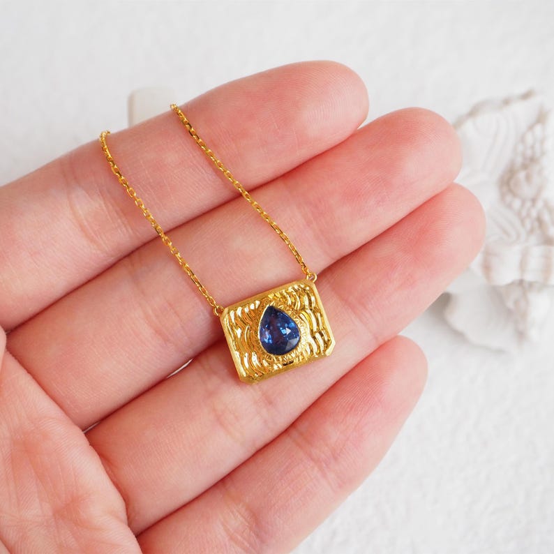 May include: A gold necklace with a rectangular pendant featuring a blue teardrop-shaped gemstone.