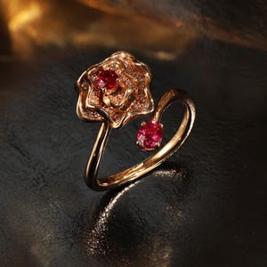 以下が含まれることがあります： 金色のリングで、花のモチーフが施されています。リングは、赤い宝石を中央に配した花型のトップと、下にぶら下がっている小さな赤い宝石が特徴です。リングは暗い背景にセットされています。