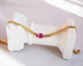 May include: A gold chain bracelet with a pink oval gemstone in the center.
