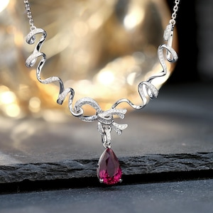 May include: A silver necklace with a bow design and a teardrop-shaped, deep red gemstone pendant. The necklace features a delicate chain and is adorned with small, sparkling stones throughout the bow and swirling accents.
