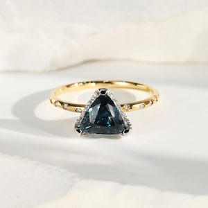 May include: A gold ring with a triangular, teal-colored gemstone. The gemstone is surrounded by small, clear stones. The band is gold with small, clear stones set into it. The ring is set against a white background.