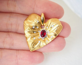 May include: A gold heart-shaped pendant with a red gemstone in the center and small white gemstones around it. The pendant has a detailed, textured design.