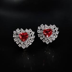 Puede incluir: Un par de pendientes de botón en forma de corazón. Cada pendiente presenta una vibrante gema roja en forma de corazón, rodeada por un halo de gemas claras más pequeñas. Los pendientes están sobre un fondo oscuro.