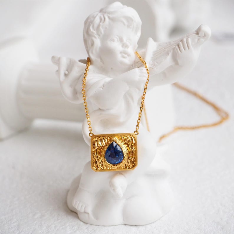 May include: Gold necklace with a rectangular pendant featuring a dark blue pear-shaped gemstone. The pendant has an ornate, textured design. The necklace is displayed on a white cherubic figurine holding a violin.