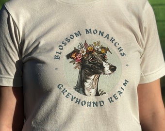 May include: Cream-colored t-shirt featuring a circular graphic with a greyhound wearing a floral crown. The text "BLOSSOM MONARCHS GREYHOUND REALM" is printed around the image in a teal color. The shirt is made of a soft material.
