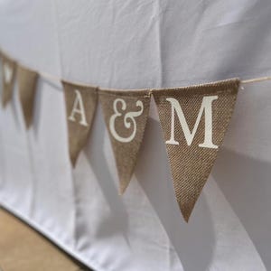 Könnte beinhalten: Ein Wimpelbanner aus Jute mit weißen Buchstaben "A & M" hängt vor einer weißen Oberfläche. Das Banner besteht aus kleinen, dreieckigen Flaggen, die aneinandergereiht sind. Der Hintergrund ist unscharf, was auf eine Umgebung im Freien hindeutet.