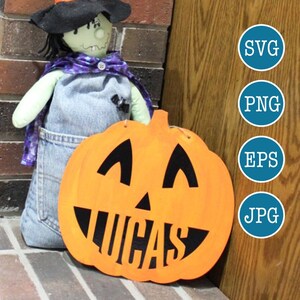 May include: A Halloween-themed pumpkin with a jack-o'-lantern face and the name "Lucas" cut out. The pumpkin is orange with black features and is next to a witch doll with a purple and black cape.