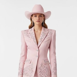 May include: A light pink Western-style suit with intricate silver embroidery. The ensemble includes a blazer, matching pants, a cowboy hat, boots, and a small clutch. The suit is a monochromatic design, perfect for a themed event.