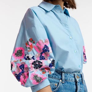 Puede incluir: Camisa azul claro con adornos florales de lentejuelas en las mangas. Las lentejuelas son rosas, naranjas y azul marino. La camisa se combina con vaqueros azules y una gorra de béisbol verde.