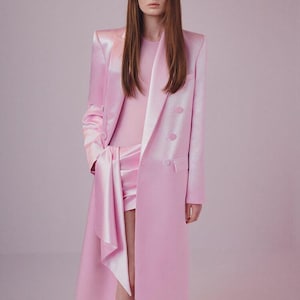 Może przedstawiać: Monochromatyczny różowy zestaw składający się z długiego płaszcza z dwurzędowym zapięciem, pasującego topu i szortów. Strój uzupełniają białe szpilki z czubkiem. Płaszcz ma asymetryczny dół.