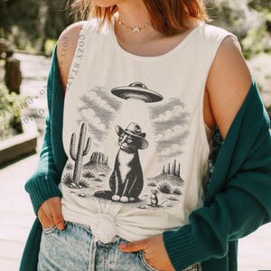 K&ouml;nnte beinhalten: Ein &auml;rmelloses, elfenbeinfarbenes Tanktop mit einem schwarzen Grafikdruck einer Katze mit Cowboyhut unter einem UFO in einer W&uuml;stenlandschaft mit Kaktus und kleiner Maus. Das Oberteil ist in der Taille geknotet und wird zu blauen Jeans getragen.