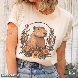 May include: Soft cream-colored t-shirt featuring a detailed illustration of a capybara surrounded by flowers within a circular frame. The shirt has short sleeves and the text "SOFT CREAM" is at the bottom. The brand name "Cozy State" is also visible.