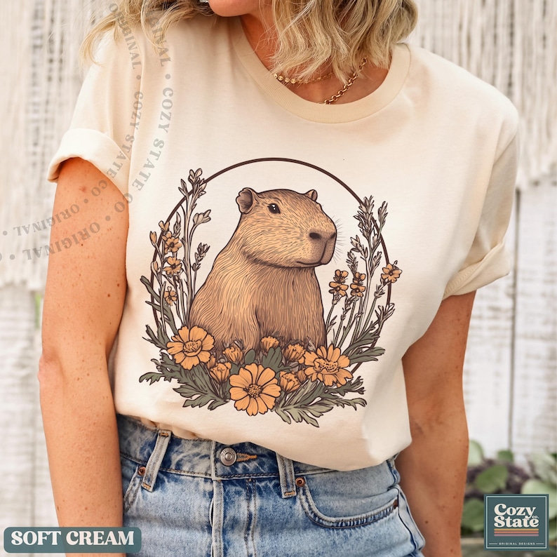May include: Soft cream-colored t-shirt featuring a detailed illustration of a capybara surrounded by flowers within a circular frame. The shirt has short sleeves and the text "SOFT CREAM" is at the bottom. The brand name "Cozy State" is also visible.