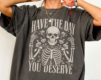 Può includere: Una t-shirt a maniche corte grigio antracite con una stampa grafica di uno scheletro con accenti floreali. Il testo "HAVE THE DAY YOU DESERVE" è stampato con un carattere chiaro sopra e sotto l'illustrazione dello scheletro sul davanti.