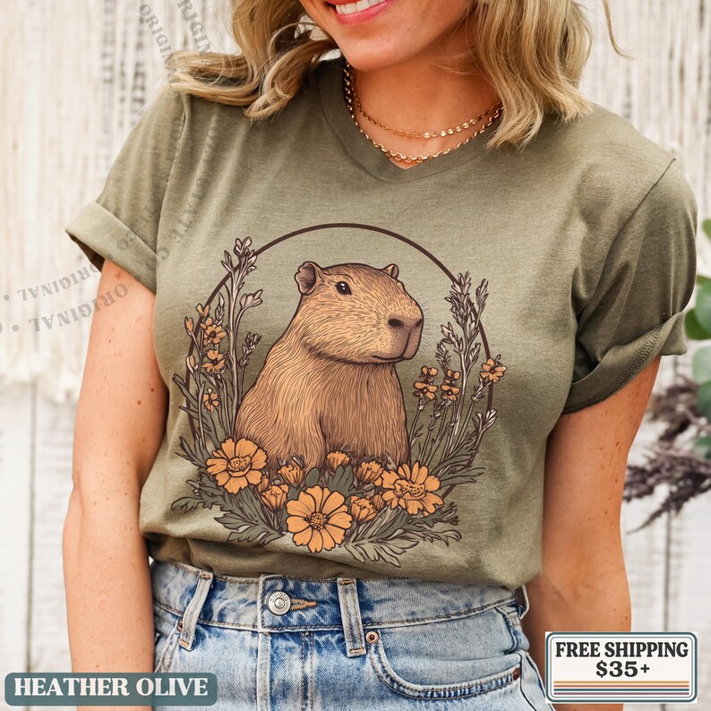 May include: Heather olive green t-shirt featuring a capybara illustration within a floral wreath. The capybara is brown, and the flowers are orange. The text "HEATHER OLIVE" is at the bottom of the shirt.