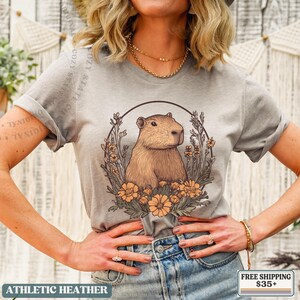 May include: Athletic heather gray t-shirt featuring a capybara illustration within a floral wreath. The capybara is brown, and the flowers are orange. The text "COZY STATE CO" is visible. The shirt also has the text "ATHLETIC HEATHER".