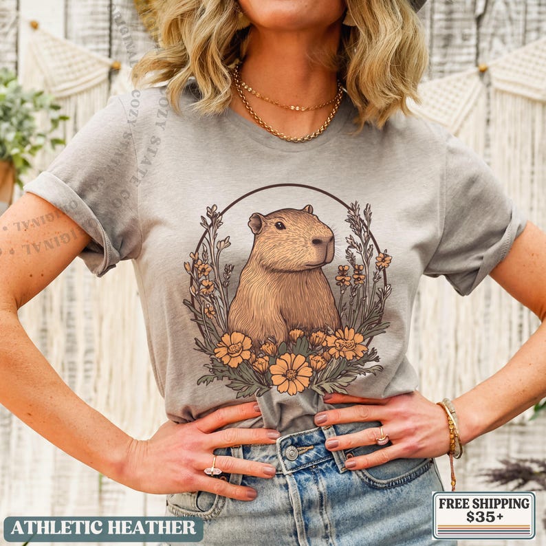 May include: Athletic heather gray t-shirt featuring a capybara illustration within a floral wreath. The capybara is brown, and the flowers are orange. The text "COZY STATE CO" is visible. The shirt also has the text "ATHLETIC HEATHER".