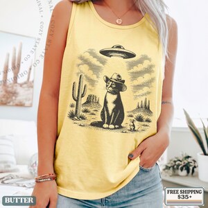 K&ouml;nnte beinhalten: Ein gelbes Tanktop mit einem schwarzen Grafikdruck einer Katze mit Cowboyhut in einer W&uuml;stenlandschaft mit Kakteen und Mesas, unter einer fliegenden Untertasse, die einen Lichtstrahl aussendet, mit einer kleinen Maus in der N&auml;he.
