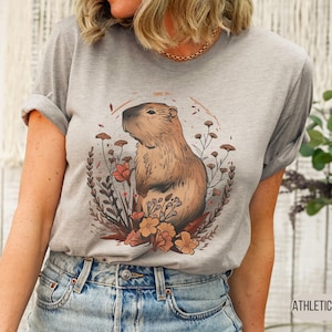May include: Athletic heather gray t-shirt featuring a capybara illustration surrounded by flowers and foliage. The capybara is brown with a detailed design. The shirt has rolled-up sleeves and the text "ATHLETIC HEATHER" is visible.