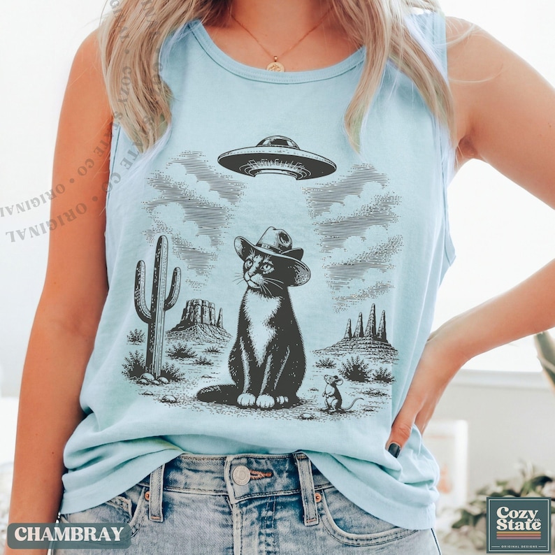 K&ouml;nnte beinhalten: Ein hellblaues &auml;rmelloses Tanktop mit einem schwarzen Grafikdruck einer Katze mit Cowboyhut in einer W&uuml;stenlandschaft. &Uuml;ber der Katze schwebt eine fliegende Untertasse am Himmel, w&auml;hrend eine kleine Maus in der N&auml;he steht. Ein Kaktus ist abgebildet.