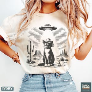 K&ouml;nnte beinhalten: Ein elfenbeinfarbenes T-Shirt mit einem schwarz-wei&szlig;en Grafikdruck einer Katze mit Cowboyhut in einer W&uuml;stenlandschaft. &Uuml;ber der Katze strahlt eine fliegende Untertasse einen Lichtstrahl aus. Ein Kaktus, Felsformationen und eine kleine Maus sind ebenfalls abgebildet.