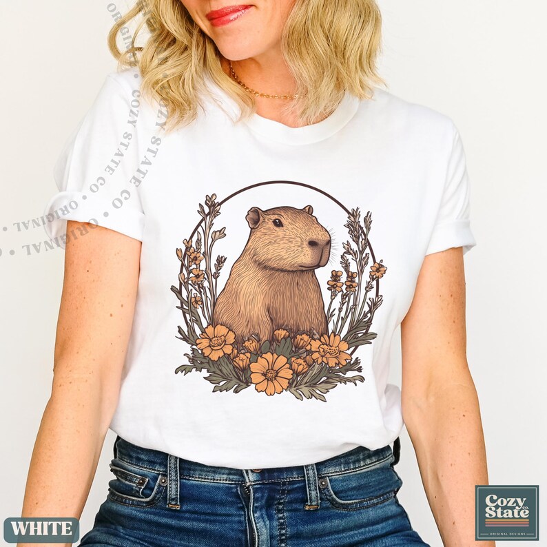 May include: White t-shirt featuring a capybara illustration within a circular floral design. The capybara is depicted in shades of brown, surrounded by orange flowers and green foliage. The shirt has a crew neck and short sleeves.